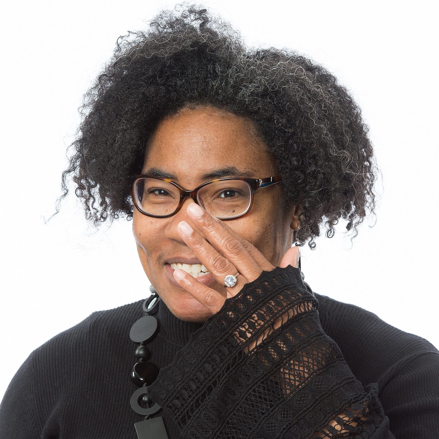 A headshot of DeMisty D. Bellinger, a Black woman with glasses smiling at the camera as she touches her left hand to her face.