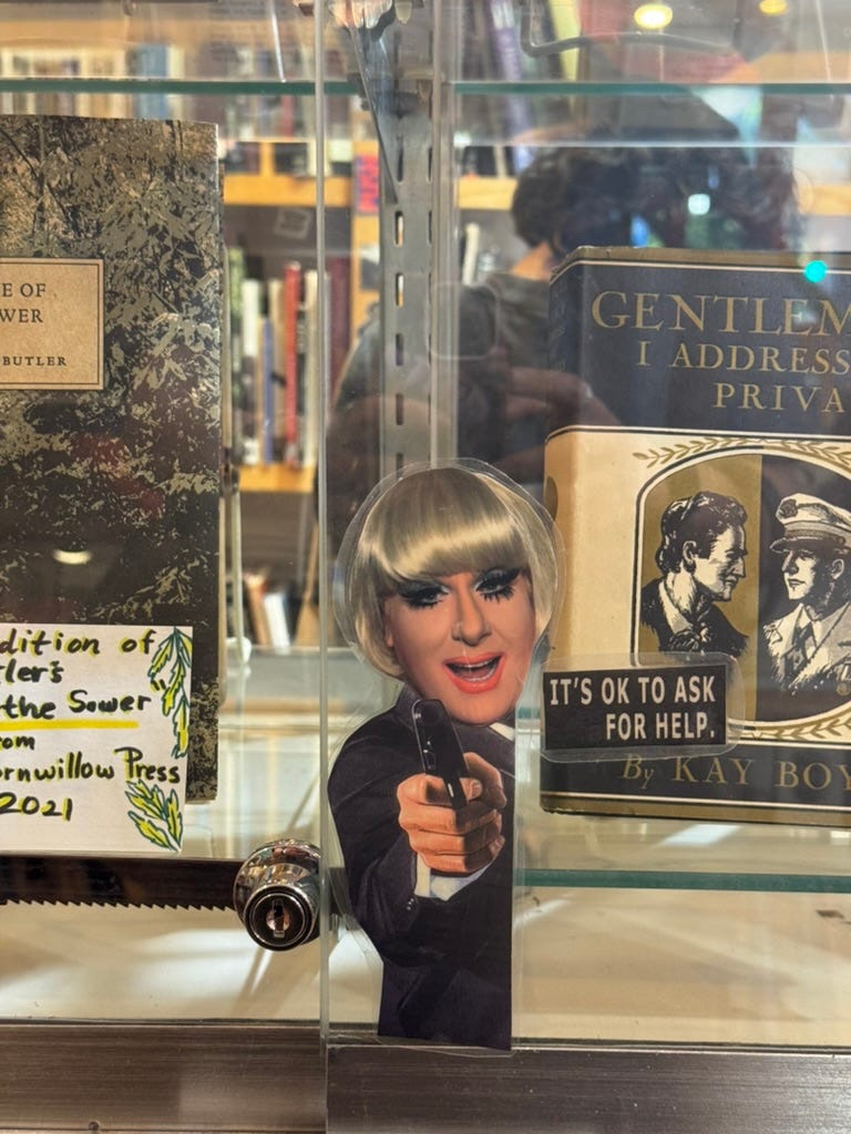 A picture Kat took at Philly AIDS Thrift @ Giovanni’s Room The Aids Thrift At Giovanni’s Room. A photo of Lady Bunny pointing a gun is taped onto the glass of a book display with the words “it’s ok to ask for help” taped beside her. 