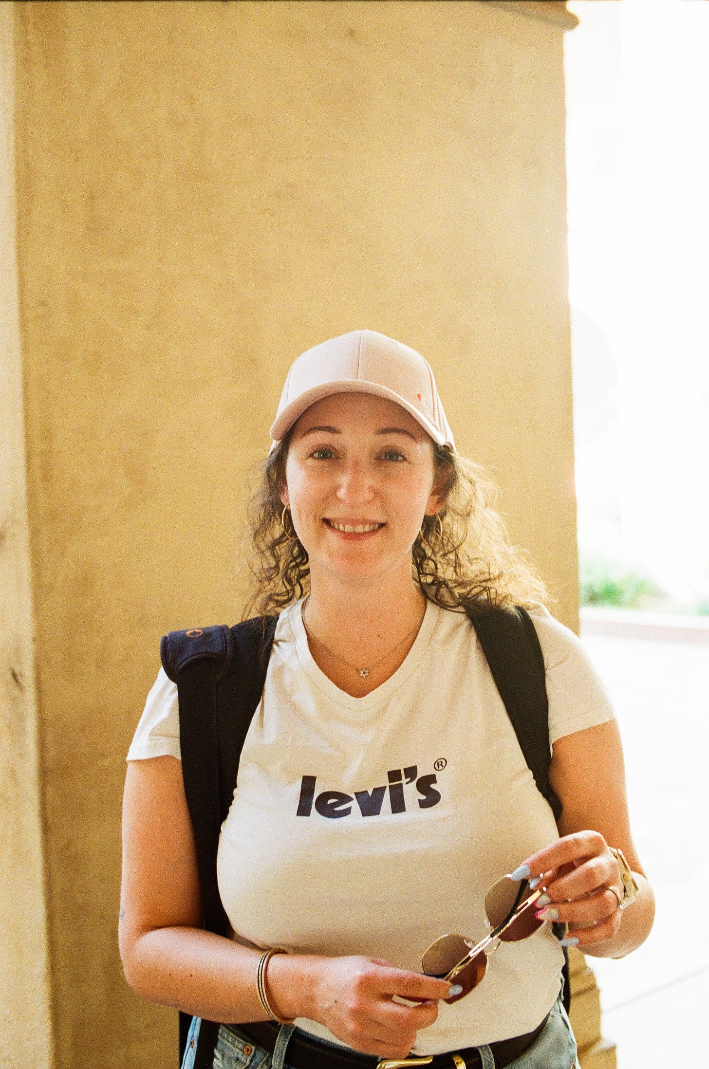 Woman with backpack, hat, and sunglasses smiling in a shaded courtyard area of Balboa Park