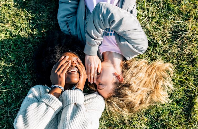 Two smiling people lying on grass, one with hands over eyes and the other whispering into their ear Two smiling people lying on grass, one with hands over eyes and the other whispering into their ear
