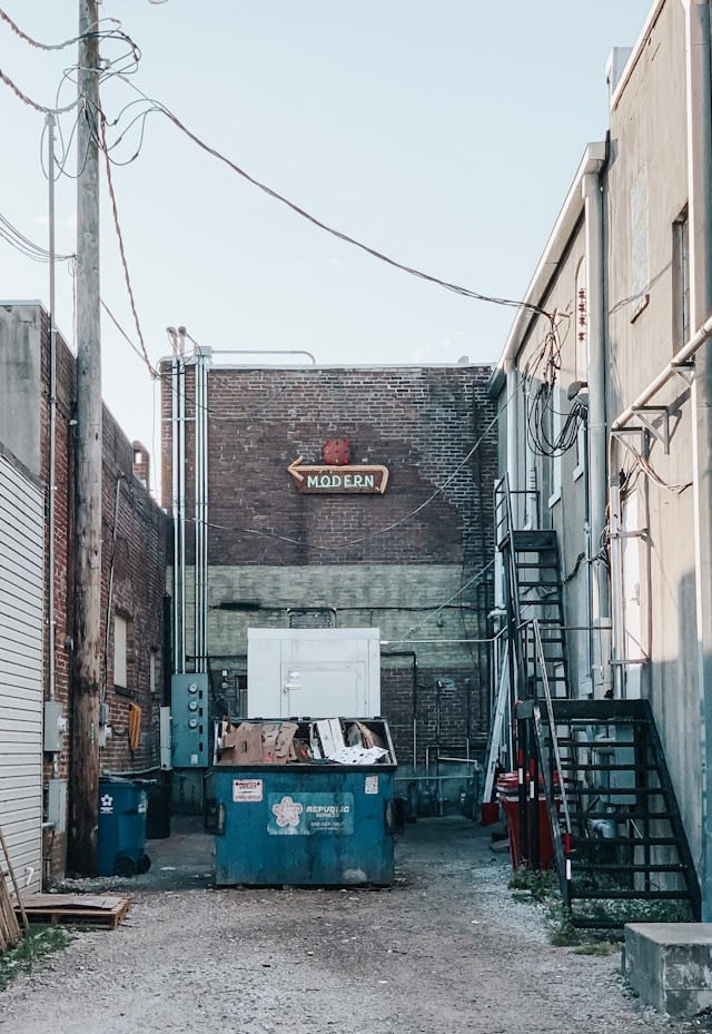A blue dumpster is overflowing with trash in an alleyway. Behind the dumpter is a brick wall and there is a neon sign on the wall that says "Modern" and points towards the street. 