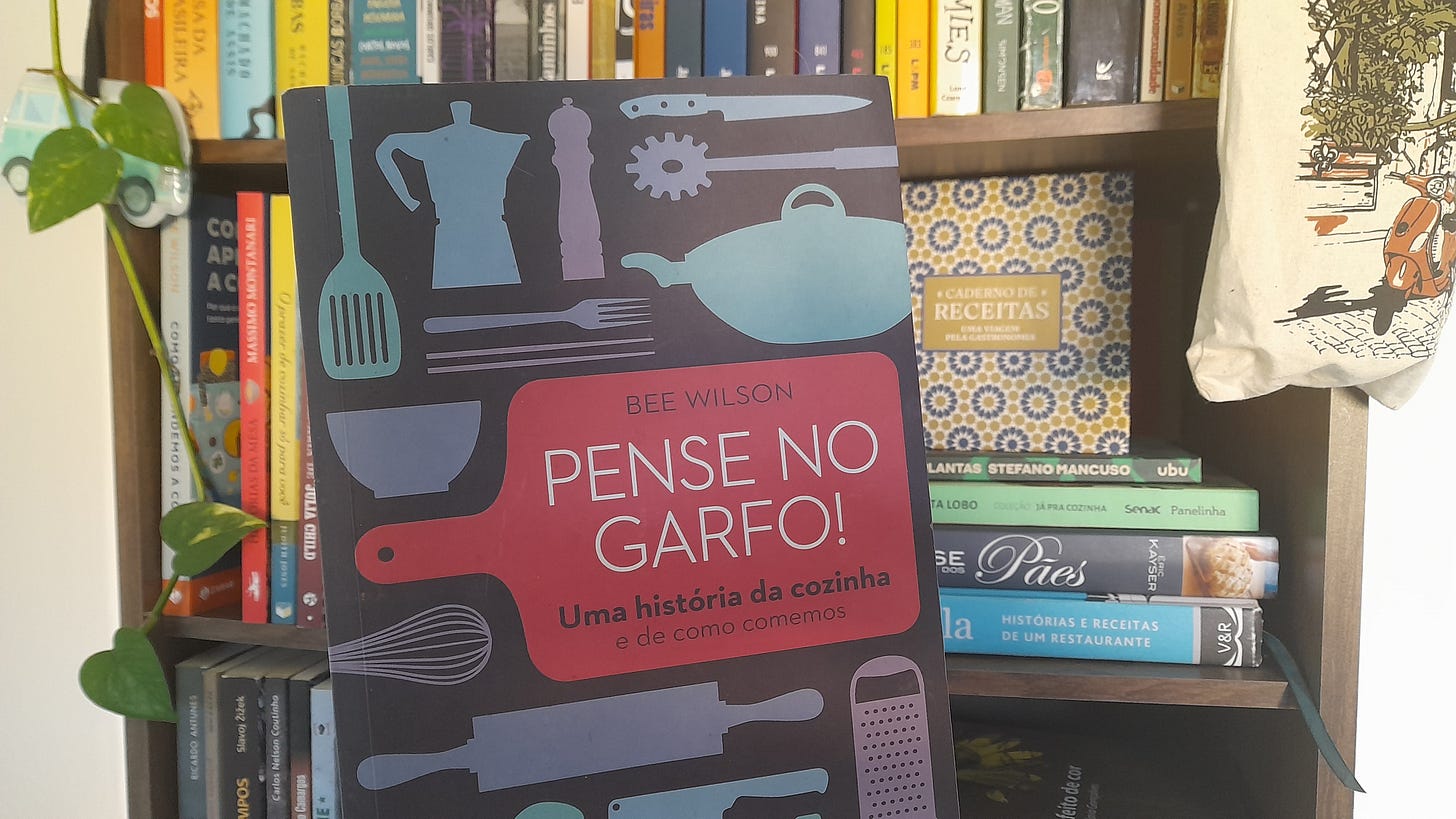 Livro "Pense no garfo", de Bee Wilson, Capa azul-escuro com figuras de utensílios de cozinha em outros tons de azul. O título está escrito em branco sobre a figura de uma tábua de corte cor-de-rosa. Ao fundo, uma estante de livros com uma planta pendente à esquerda e uma ecobag à direita. Livro "Pense no garfo", de Bee Wilson, Capa azul-escuro com figuras de utensílios de cozinha em outros tons de azul. O título está escrito em branco sobre a figura de uma tábua de corte cor-de-rosa. Ao fundo, uma estante de livros com uma planta pendente à esquerda e uma ecobag à direita.