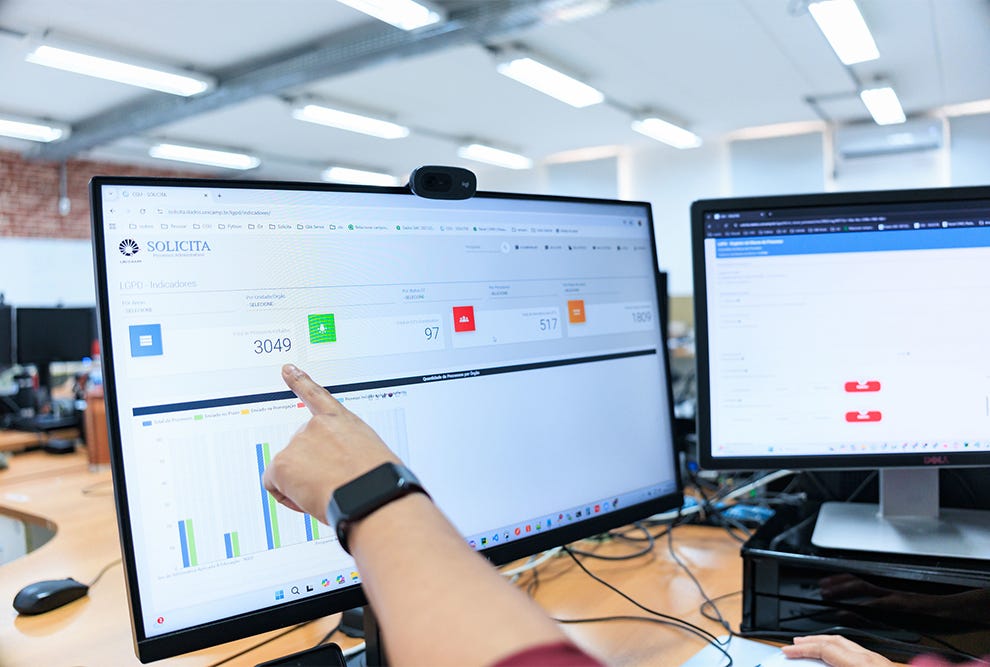 Pessoa com pulseira preta no pulso esquerdo apontando para monitor de computador exibindo dashboard com logo Solicita, números em cards coloridos (3049, 97, 517), gráficos de barras em azul e verde, em ambiente de escritório com teto branco iluminado por painéis de LED, segundo monitor ao fundo à direita com interface de código, mouse e teclado sobre mesa de madeira clara. Pessoa com pulseira preta no pulso esquerdo apontando para monitor de computador exibindo dashboard com logo Solicita, números em cards coloridos (3049, 97, 517), gráficos de barras em azul e verde, em ambiente de escritório com teto branco iluminado por painéis de LED, segundo monitor ao fundo à direita com interface de código, mouse e teclado sobre mesa de madeira clara.