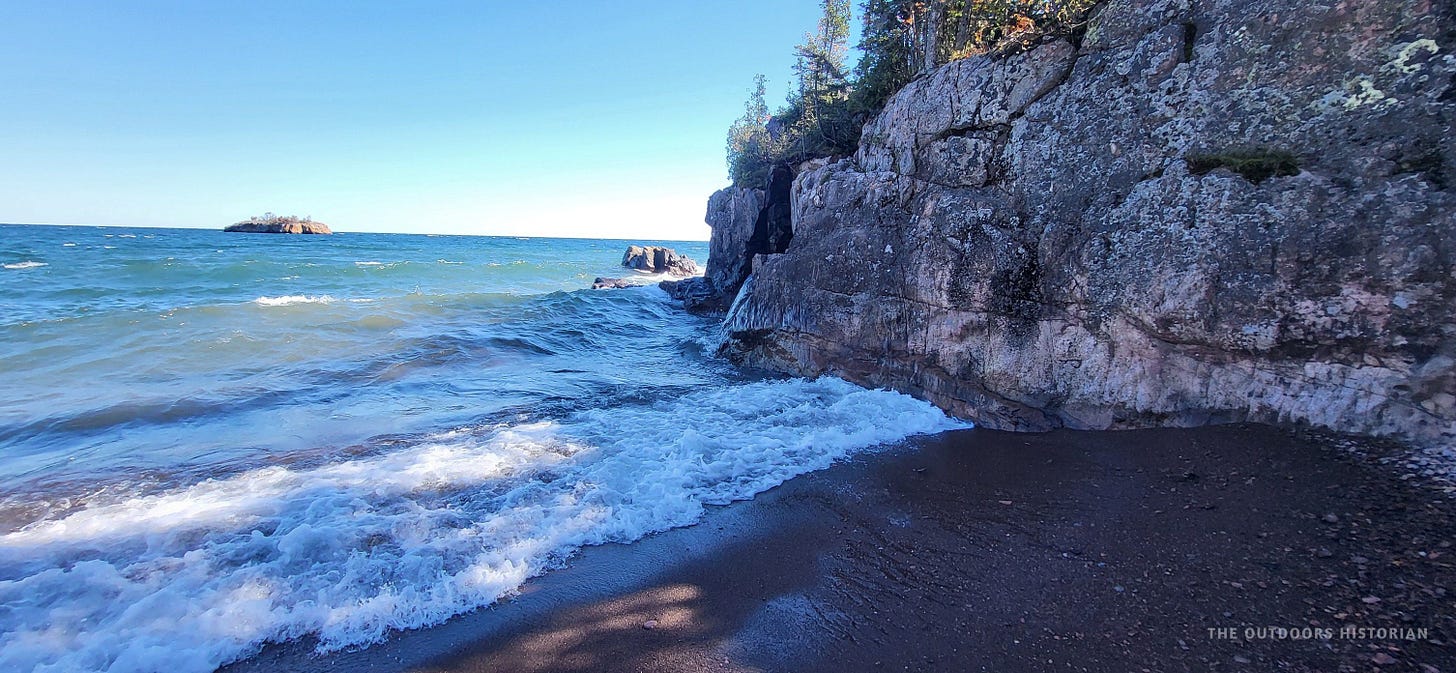 A wide shot of Black Beach in Silver Bay, Minnesota, on a sunny late September day. Dark, charcoal-colored sand is met by rhythmic, white-capped waves of Lake Superior under a clear blue sky.