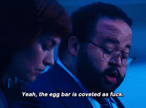 a man in a suit and tie says " yeah the egg bar is coveted as fuck " next to a woman