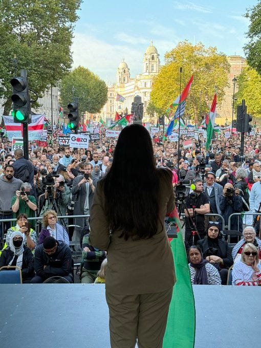 A woman with long dark hair wearing a beige blazer and trousers stands on a wooden stage facing away from the camera toward a large crowd in an urban square lined with trees showing fall colors and traffic lights. The crowd holds green Palestinian flags, red Yemeni flags, and white banners with text including FREE PALESTINE and GAZA, surrounded by photographers with cameras and diverse participants in various attire including headscarves. In the background, historic domed buildings and autumn foliage are visible under a clear sky.