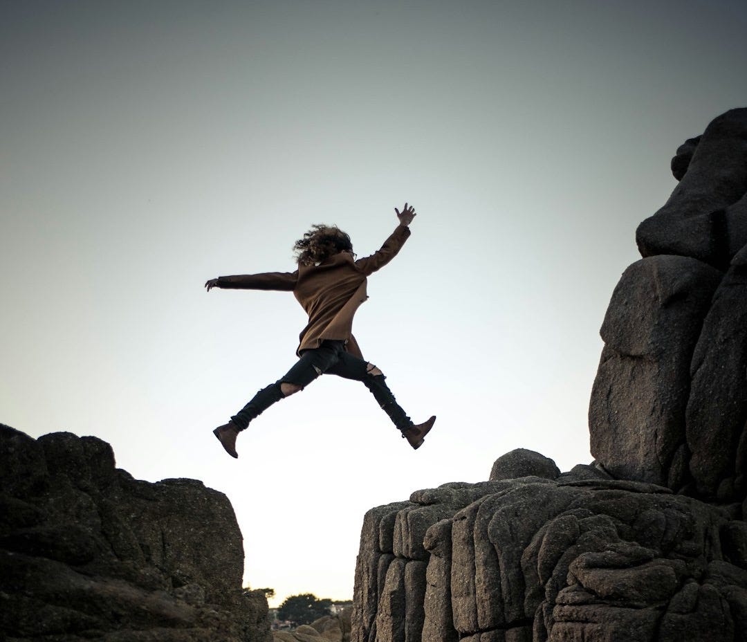 person jumping on big rock under gray and white sky during daytime