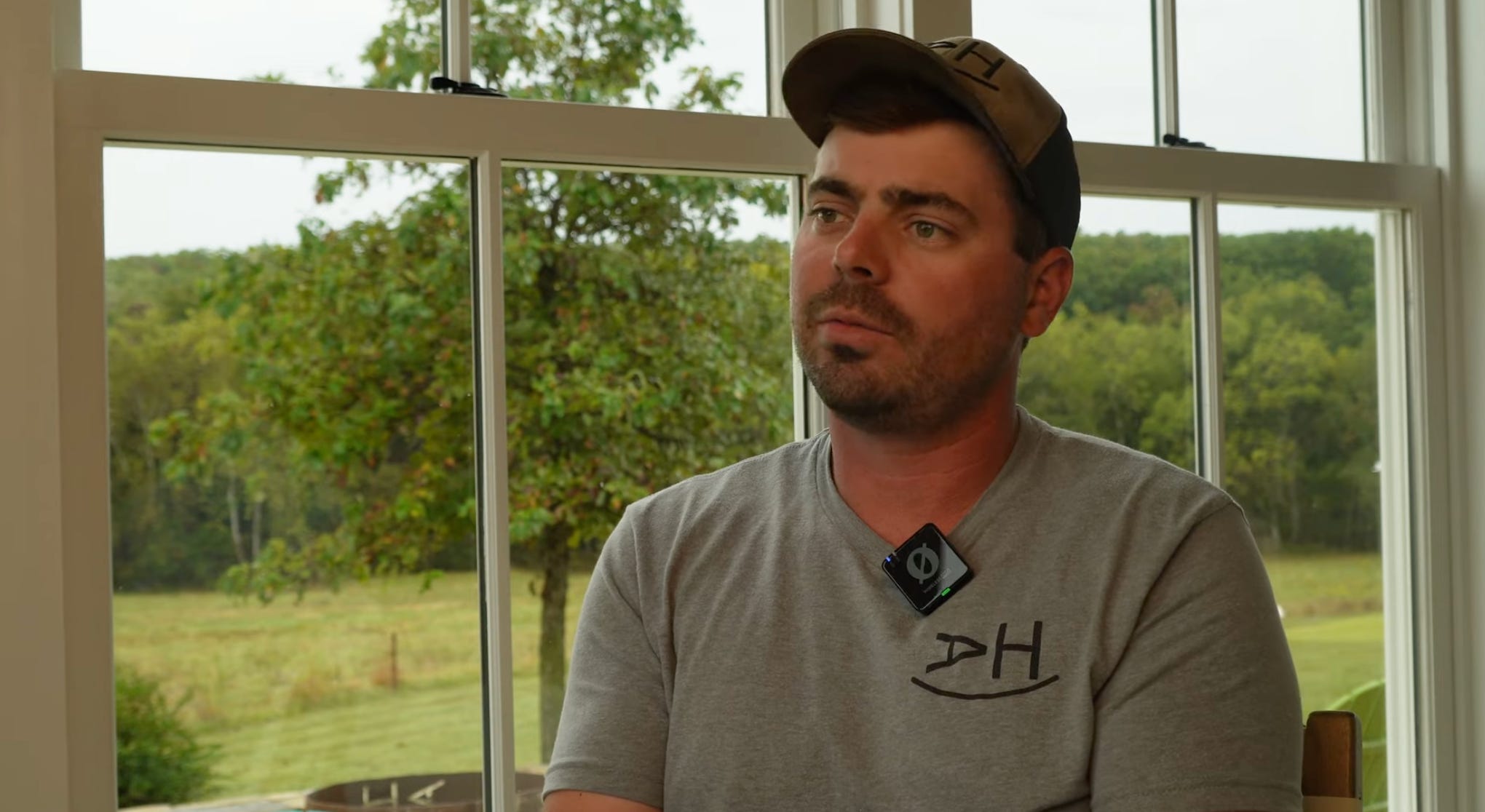 A man wearing a tan cap and gray shirt sits indoors near large windows overlooking green pastures, speaking during an interview about regenerative farming. A man wearing a tan cap and gray shirt sits indoors near large windows overlooking green pastures, speaking during an interview about regenerative farming.