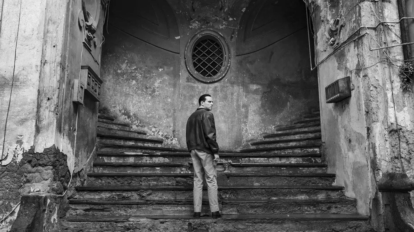 A white man standing at the base of a worn split staircase, looking back over his shoulder in an old, decaying building.