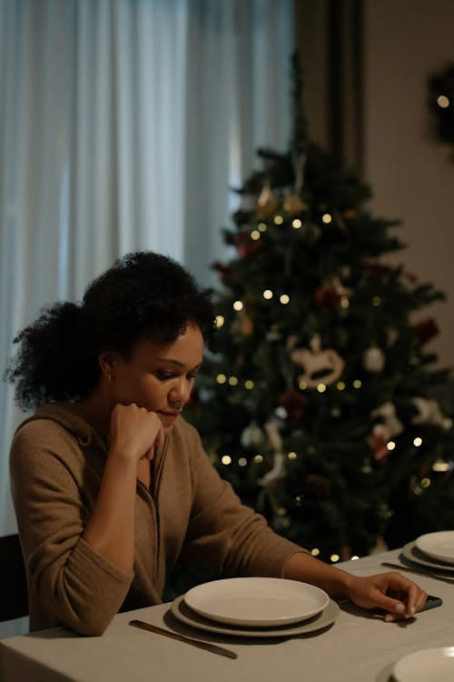 Free A solitary woman sitting at a dining table beside a decorated Christmas tree. Stock Photo