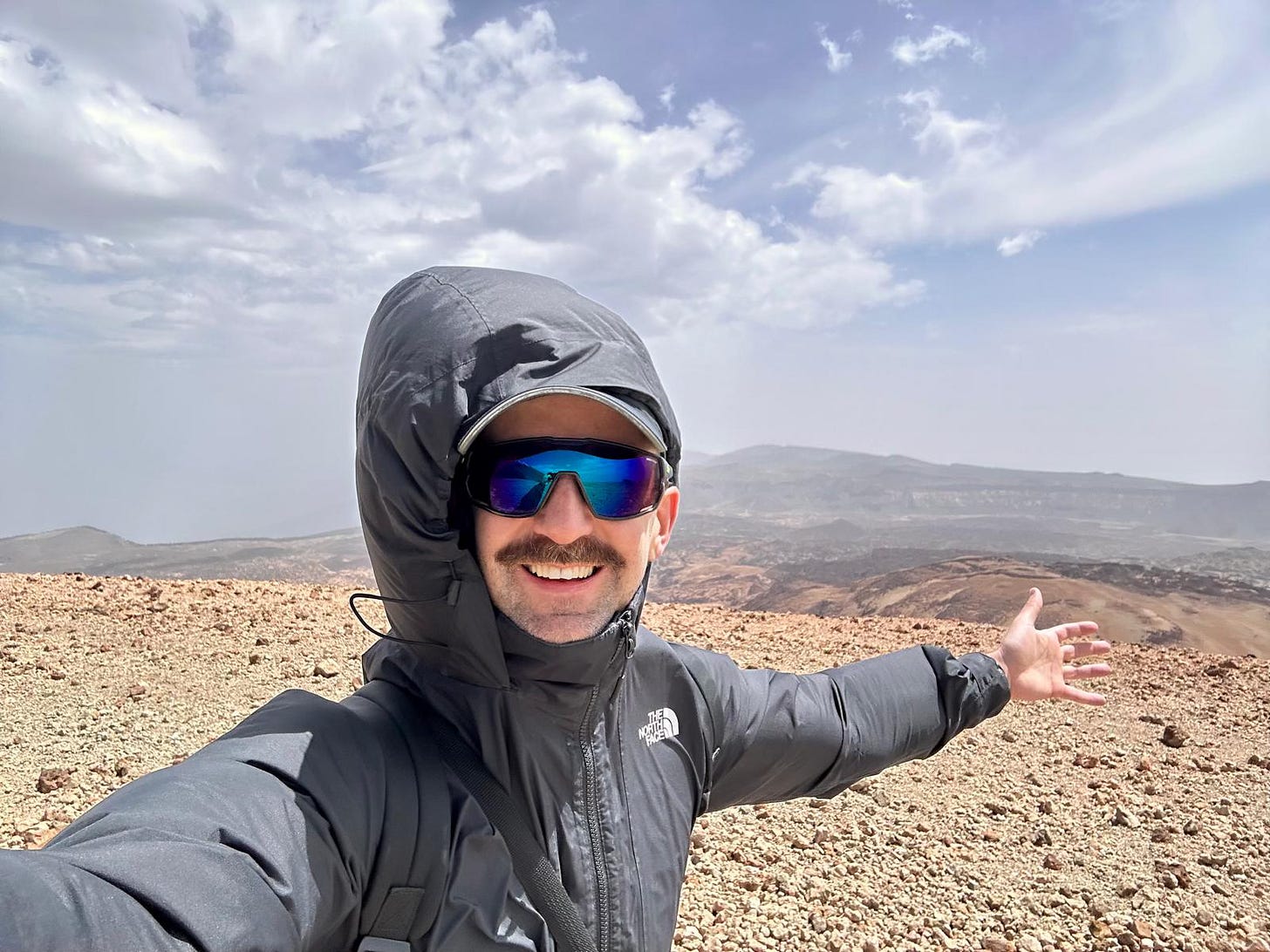 A person standing alone at the end of the hiking route at Montana Blanca, smiling widely with one hand open gesturing toward the vast volcanic landscape behind them
