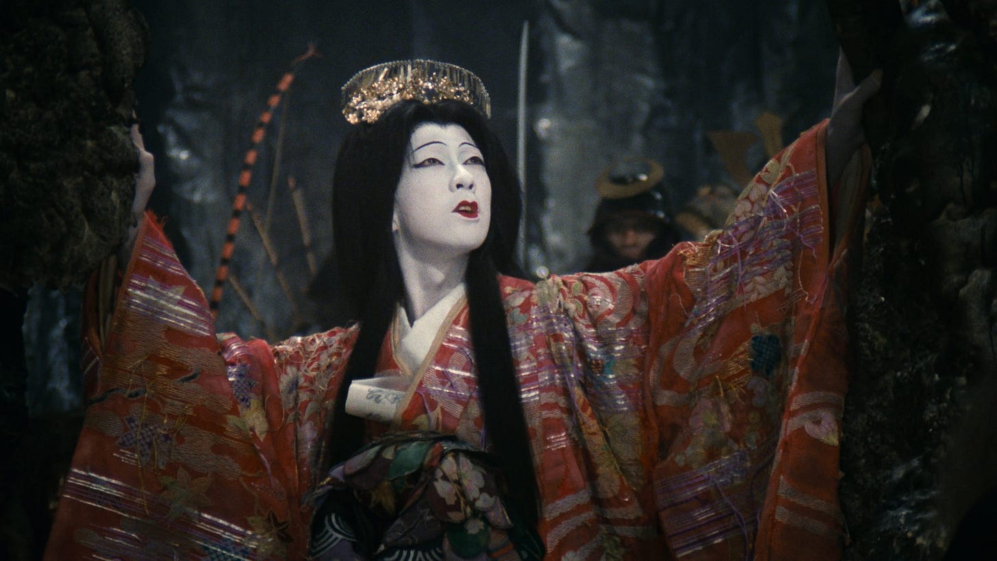 Film still from 1979 movie Demon Pond. A Japanese woman with white makeup, thin eyebrows, and bold red lips stares into the distance. She’s wearing an ornate round gold headpiece, and her striking red floral kimono is adorned with some thin silver stripes. The background is dark and ominous.