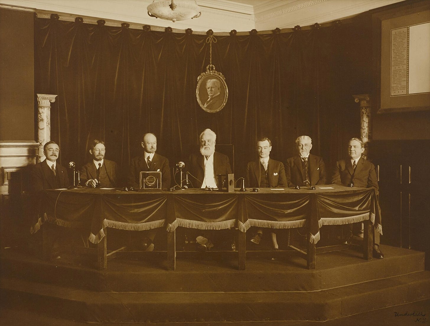 Sepia-toned photograph of Alexander Graham Bell seated at the center of a long table during the first transcontinental telephone call in 1915, with early telephone equipment, microphones, a replica of Bell’s original telephone, and other historical objects arranged in front of the participants. Sepia-toned photograph of Alexander Graham Bell seated at the center of a long table during the first transcontinental telephone call in 1915, with early telephone equipment, microphones, a replica of Bell’s original telephone, and other historical objects arranged in front of the participants.