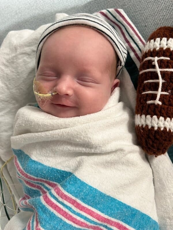 A close-up portrait of newborn KJ, who has a tube coming out of his nose and a tiny felt football next to him. He is swaddled tight and appears to smile slightly with his eyes closed. A close-up portrait of newborn KJ, who has a tube coming out of his nose and a tiny felt football next to him. He is swaddled tight and appears to smile slightly with his eyes closed.