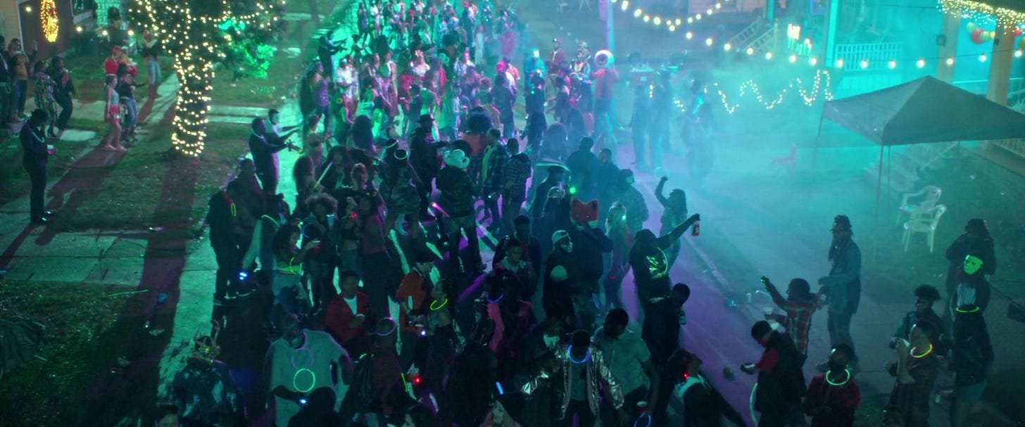 A neighborhood street is crowded with partiers carrying glowsticks. Trees, homes, and hedges are decorated with string lights.