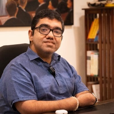 A photograph of Nipun Malhotra with short black hair, glasses, and a calm expression sitting at a desk with his arms resting on the surface. He wears a blue collared shirt and a small microphone clipped to it. A bookshelf filled with colourful books and a painting with several people in the background are visible behind him.