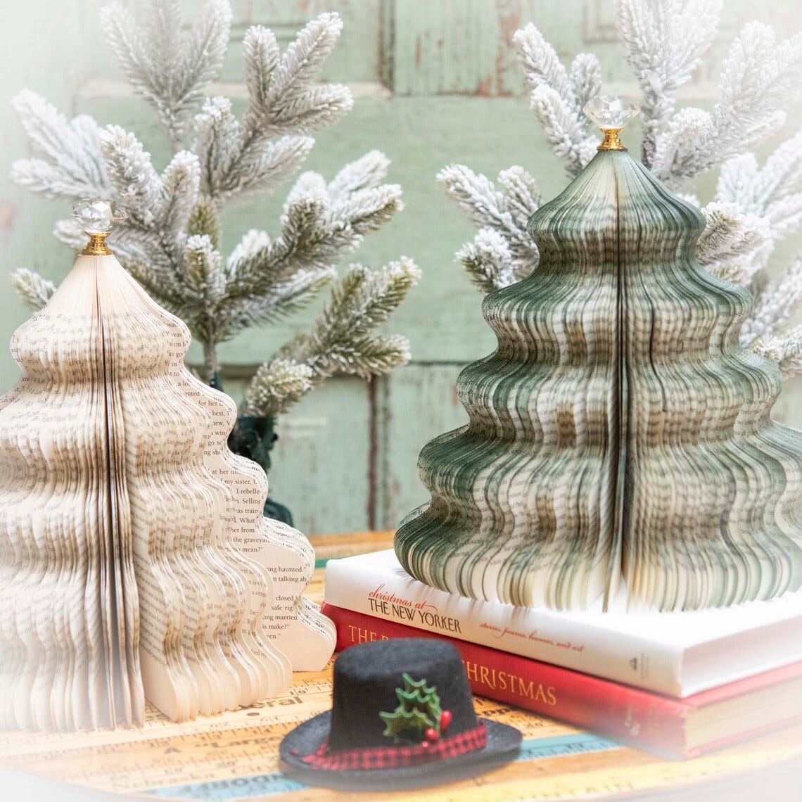 May include: Two book trees made from folded pages, one is white and the other is green. The trees are sitting on a table with a black and red hat and books stacked underneath. The books are titled 'Christmas at The New Yorker' and 'Christmas'. 