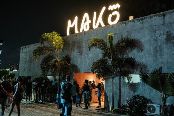 A concrete building at night with a glowing yellow "MAKŌ" sign on top. People are gathered near an orange-lit entrance, with palm trees in front. A concrete building at night with a glowing yellow "MAKŌ" sign on top. People are gathered near an orange-lit entrance, with palm trees in front.