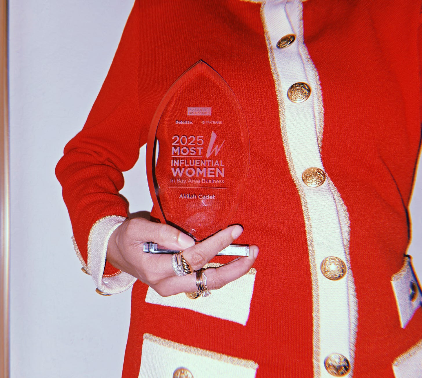 Dr. Cadet holds her glass award that says SF Business Times Most Influential Women 2025 In Bay Area Business followed by her name (without her doctor title). She wears a vintage red and cream knit with gold buttons that gives Channel vibes but it's St. John Knits.