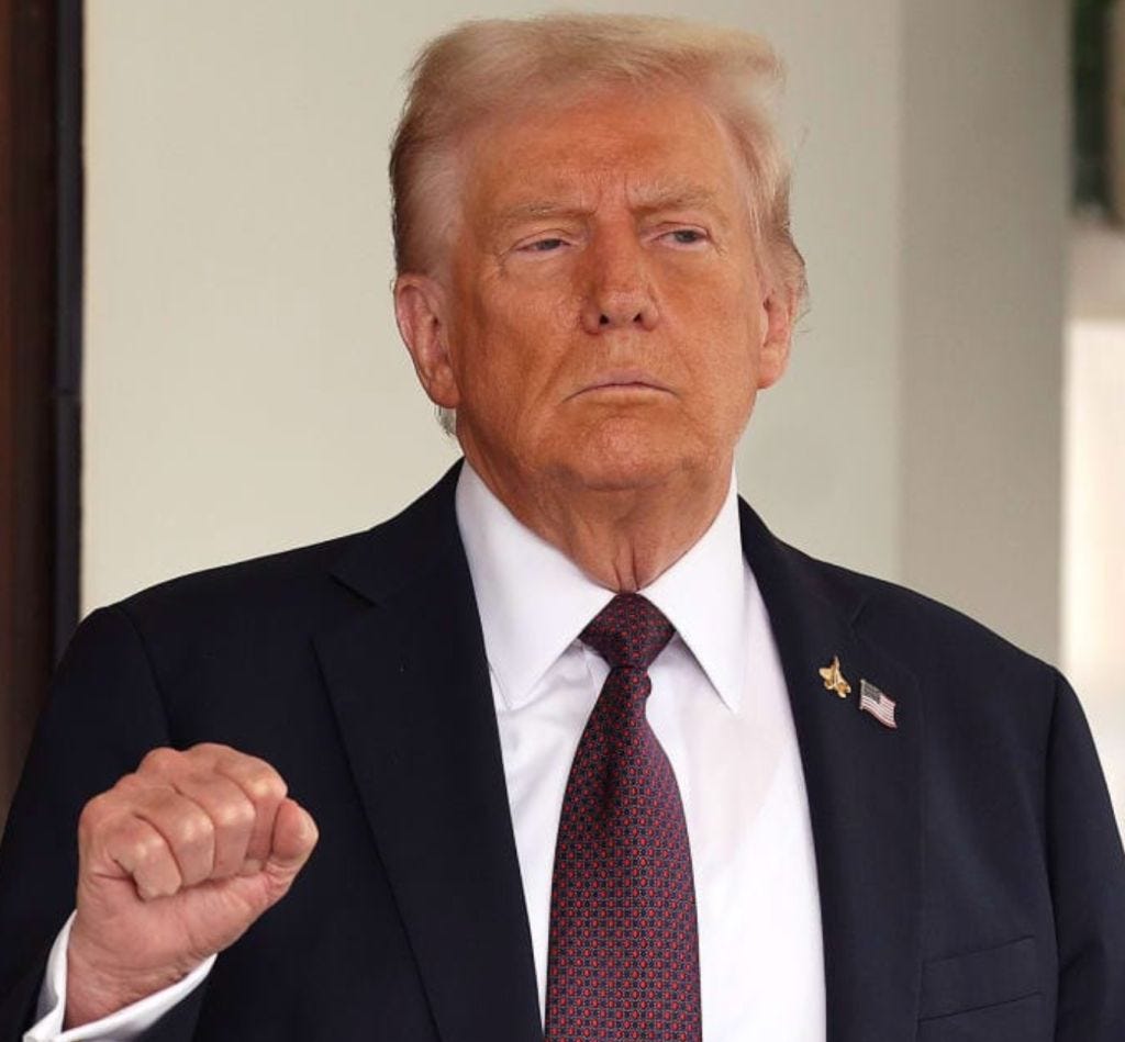 A serious-looking person with light hair and a suit is standing with a clenched fist, appearing to address an audience.