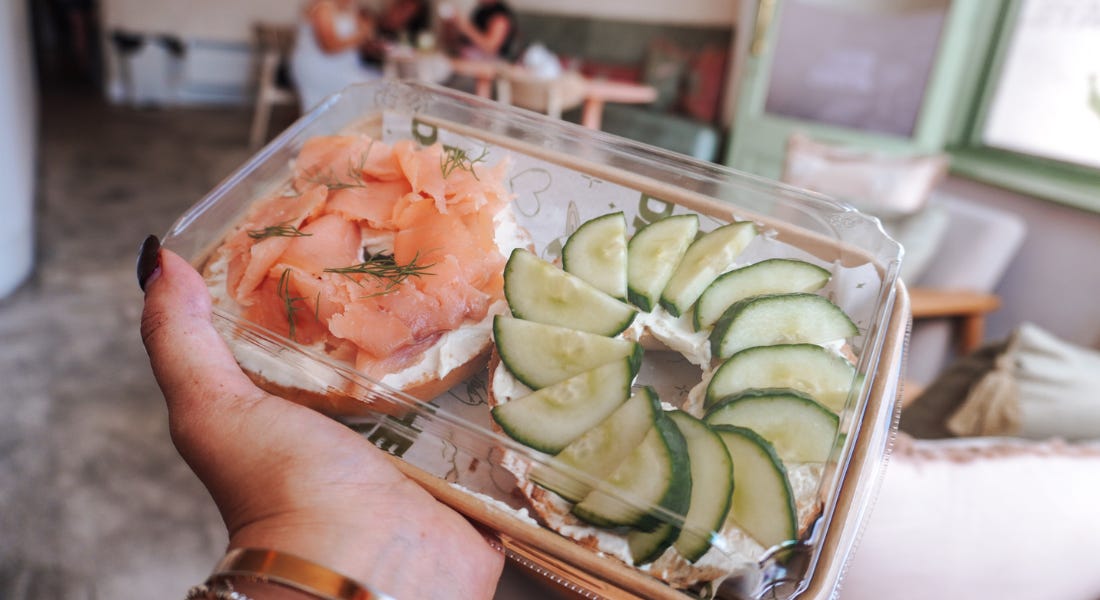 A hand holding a to-go box with a salmon and cucumber bagel in it A hand holding a to-go box with a salmon and cucumber bagel in it