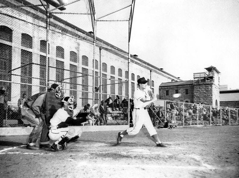 A batter swings at a pitched ball inside Folsom, with the housing block in the background.