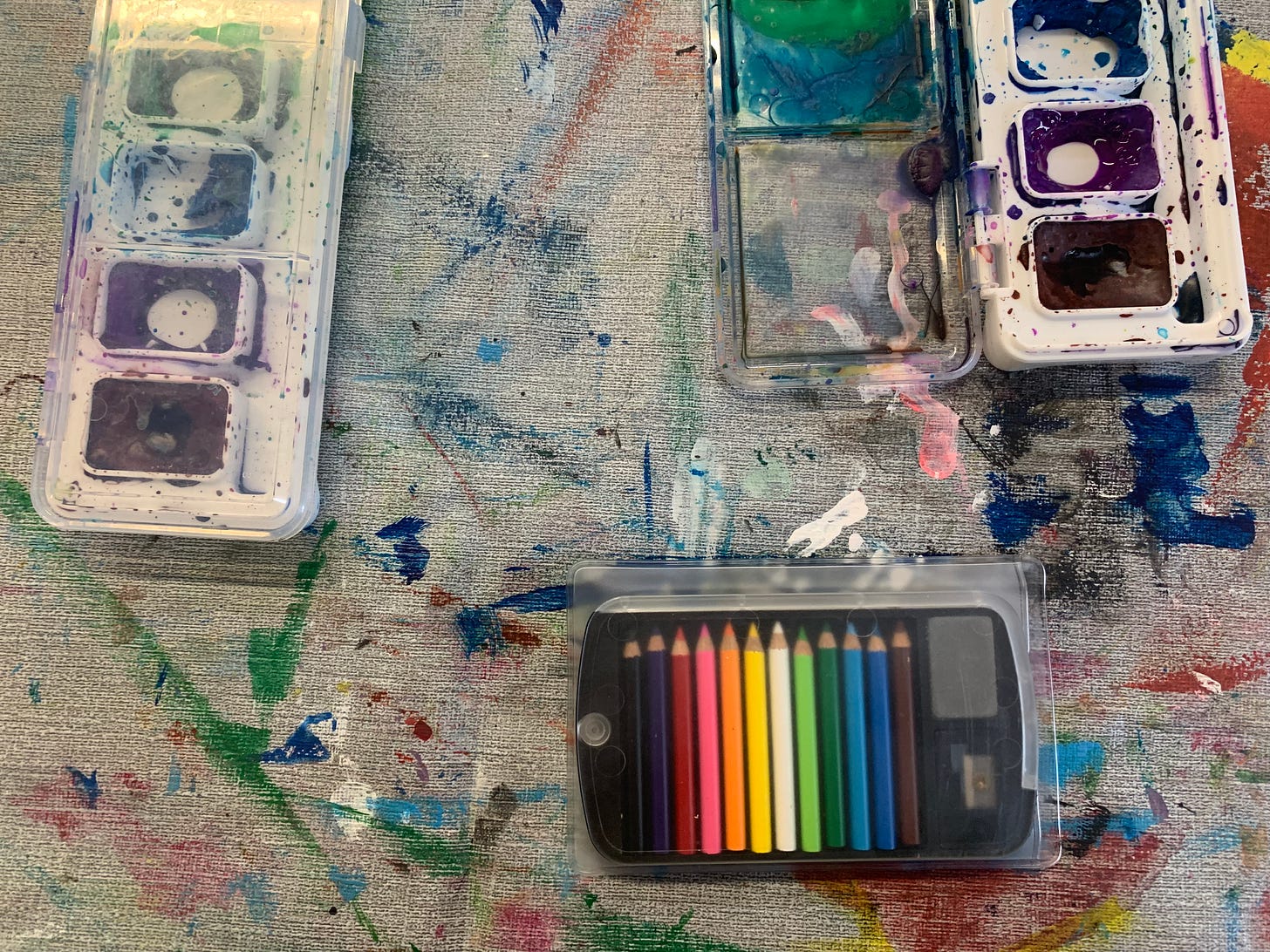 Two watercolor sets and a tiny set of colored pencils, all resting on a messy painted tablecloth