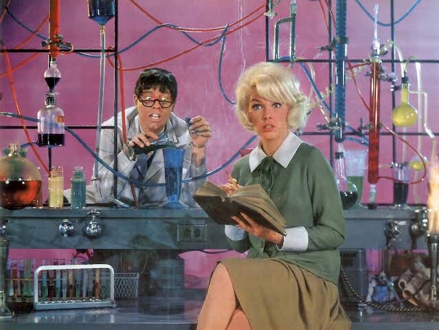 an ugly nerdy professor in background behind an apparatus of test tubes; attractive blond woman sits in front with legs crossed an ugly nerdy professor in background behind an apparatus of test tubes; attractive blond woman sits in front with legs crossed