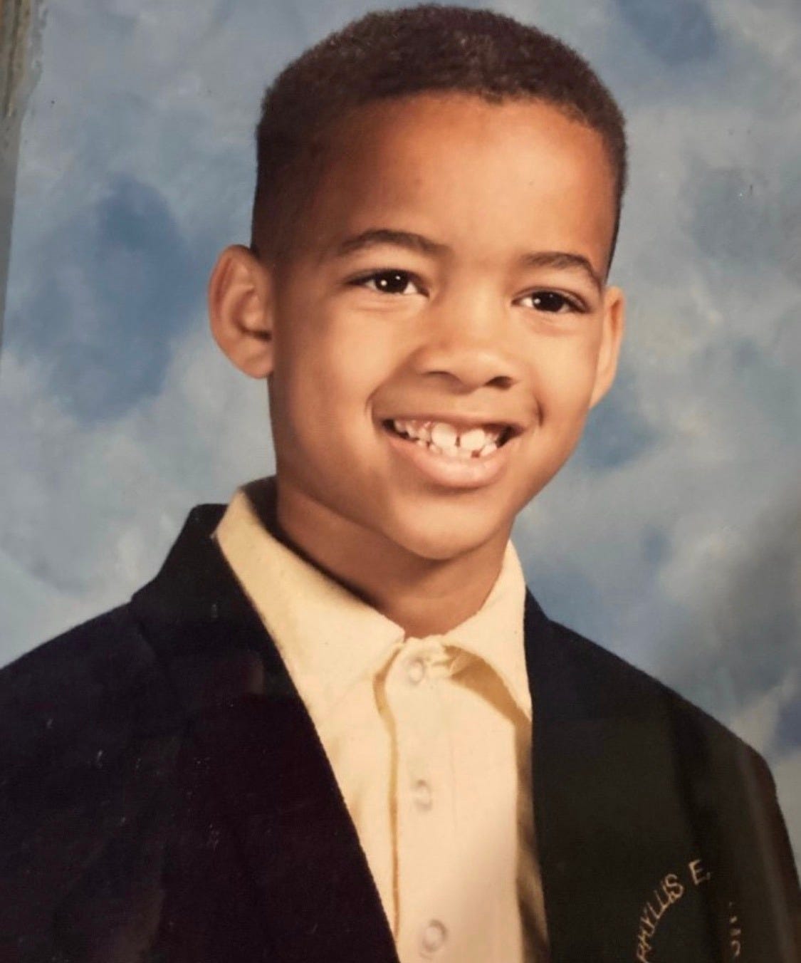 Childhood school portrait of Ethan Ward wearing a dark blazer over a light buttoned-up shirt, smiling with a slight gap in his front teeth. The background is a classic soft blue studio backdrop.