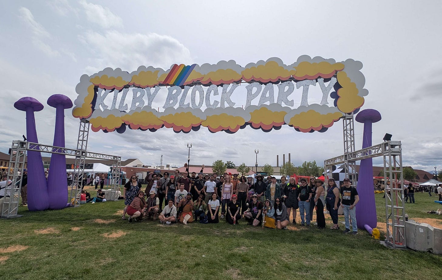 Large group of festival attendees, seated, kneeling, and standing in front of a large sign saying "KILBY BLOCK PARTY" surrounded by clouds and inflatable mushrooms.