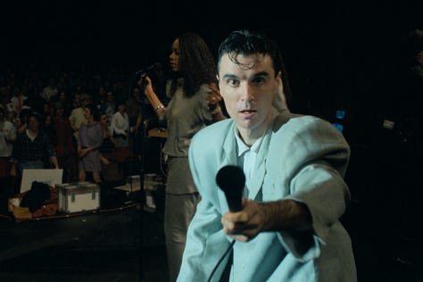 selects from Talking Heads' concert film Stop Making Sense. images of Byrne holding a microphone to camera, raising his arms, dancing in a big suit an with a lamp and full shots of the band.