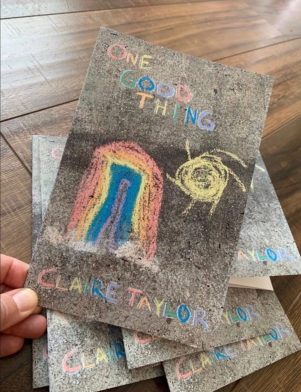 Photo of my hand holding a copy of One Good Thing which has a sidewalk chalk rainbow on the cover.