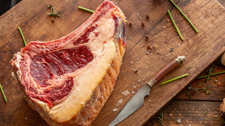 A thick raw bone-in ribeye steak rests on a wooden cutting board beside a knife, sprigs of rosemary, and salt. A thick raw bone-in ribeye steak rests on a wooden cutting board beside a knife, sprigs of rosemary, and salt.