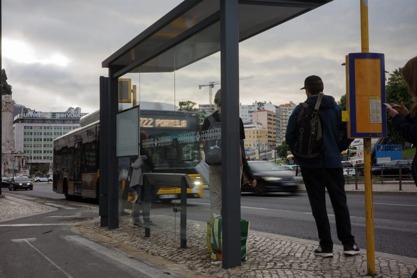 Fermata dell’autobus a Lisbona senza panchina, con persone in piedi in attesa sotto una pensilina moderna in vetro e metallo; sullo sfondo traffico urbano e palazzi residenziali.