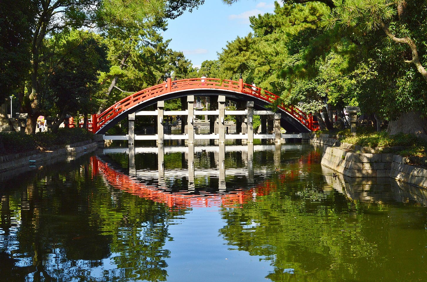 A picture of a curved bridge reflected in water at Sumiyoshi Taisha Grand Shrine, Osaka. The bridge is curved in a steep arch, has vermilion rails. Pine trees line a moat in which the bridge is reflected A picture of a curved bridge reflected in water at Sumiyoshi Taisha Grand Shrine, Osaka. The bridge is curved in a steep arch, has vermilion rails. Pine trees line a moat in which the bridge is reflected