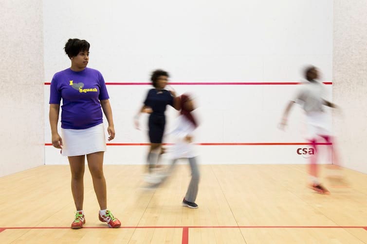 Woman in purple T-shirt and short white skirt stands on squash court as kids play