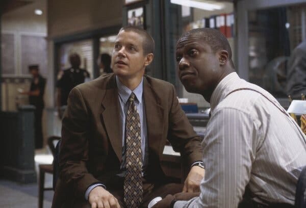 One man in a brown suit an another in a gray shirt sit together at a desk