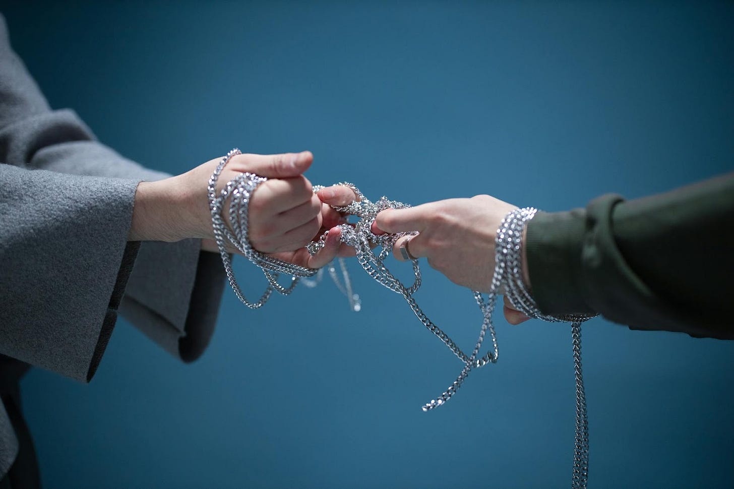 Close-up of two hands wrapped in silver chains, symbolizing connection or struggle, set against a blue backdrop.