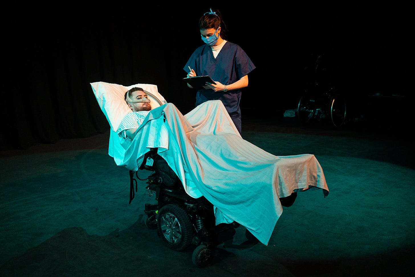 Image: Jamie Hale, a white person with dark red hair and a ginger beard lies in an electric wheelchair made up like a hospital bed. A white woman in blue scrubs and a blue facemask is looking at them, ticking something on a clipboard.