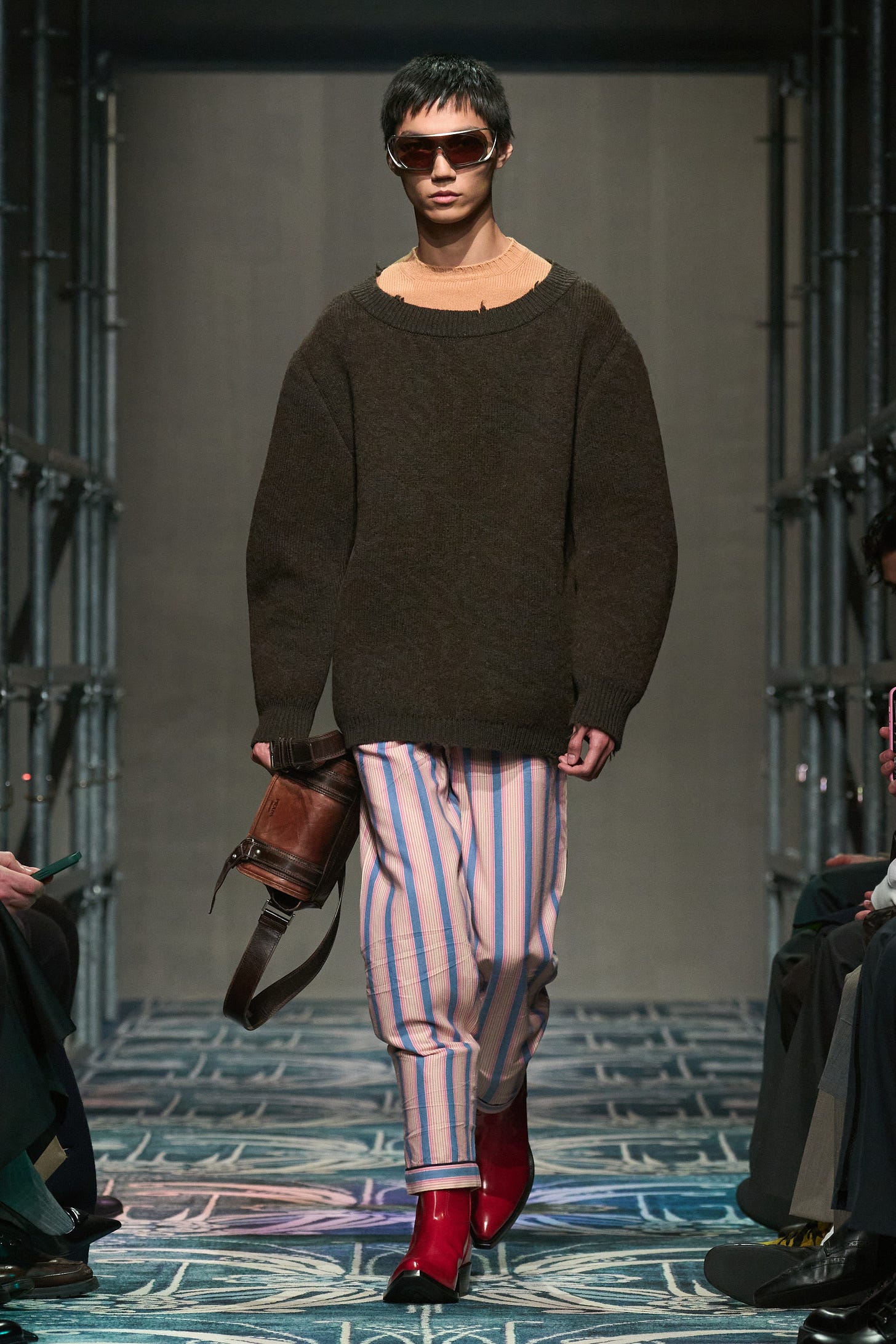 A model walks the runway wearing an oversized gray-brown sweater with striped pajama pants and cowboy boots.