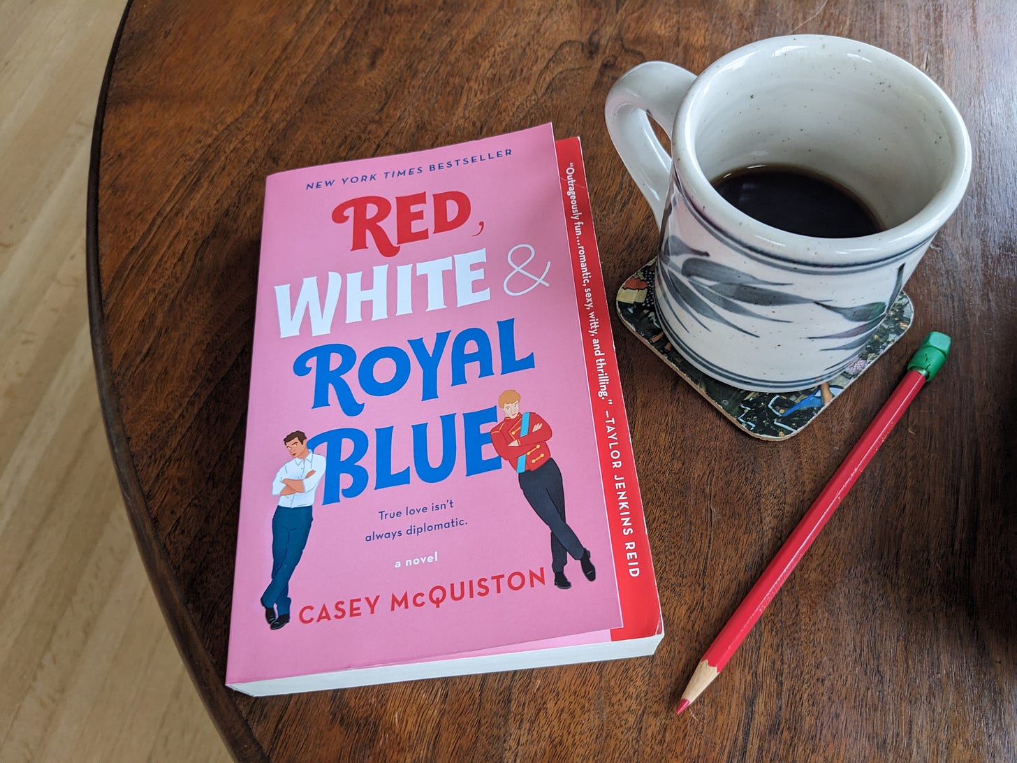 Photo of a section of a round walnut wood table on which sits a pink-covered book, a ceramic mug with a quarter of a cup of black coffee remaining, and a red colored pencil with a grass green eraser head.