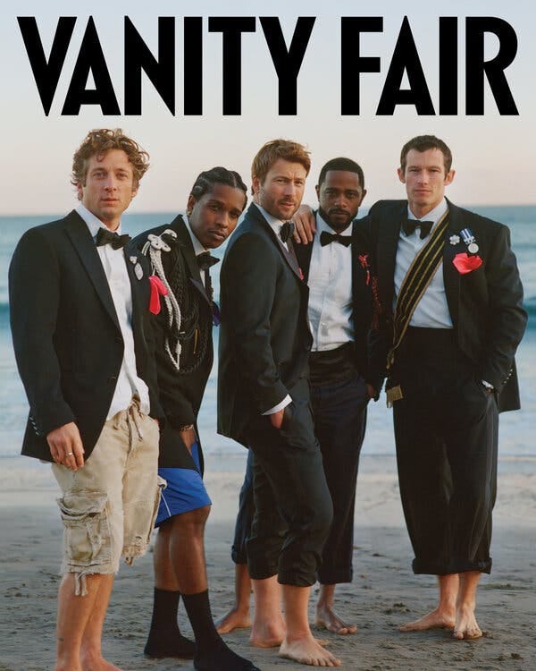 Five men in tuxedo jackets and bow ties stand on the beach. Two of the men are wearing shorts and the others have their pants rolled up.