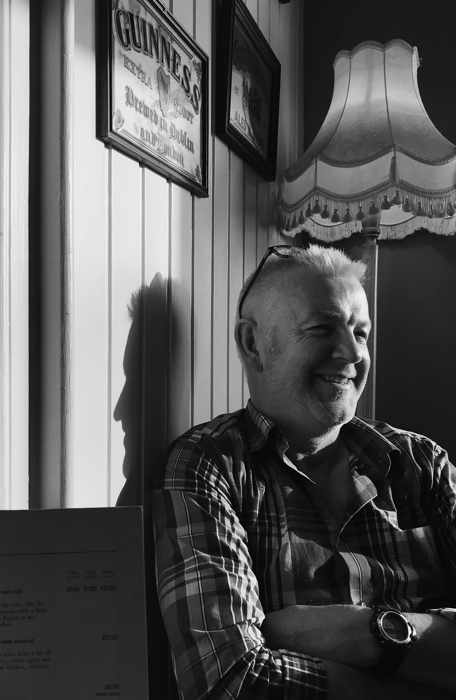 A man with white hair and glasses perched on top of his head sits smiling indoors, seated near a tasseled lampshade and a Guinness sign; his shadow can be seen next to him. A man with white hair and glasses perched on top of his head sits smiling indoors, seated near a tasseled lampshade and a Guinness sign; his shadow can be seen next to him.