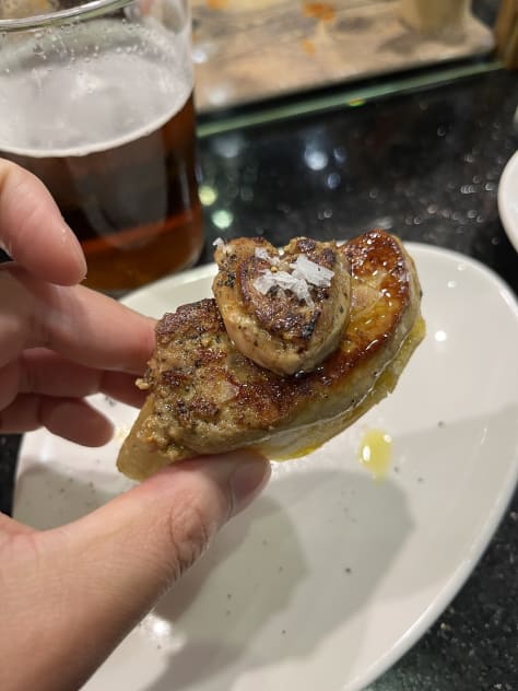 Crab stuffed squid, a spoon entering creamed uni in its shell, a hand holding a sesame crack with three different color pieces of smoked fish on top , a hand holding grilled foie on bread, a hand holding txistorra skewered onto a piece of bread, a fork entering a half eaten piece of cheesecake