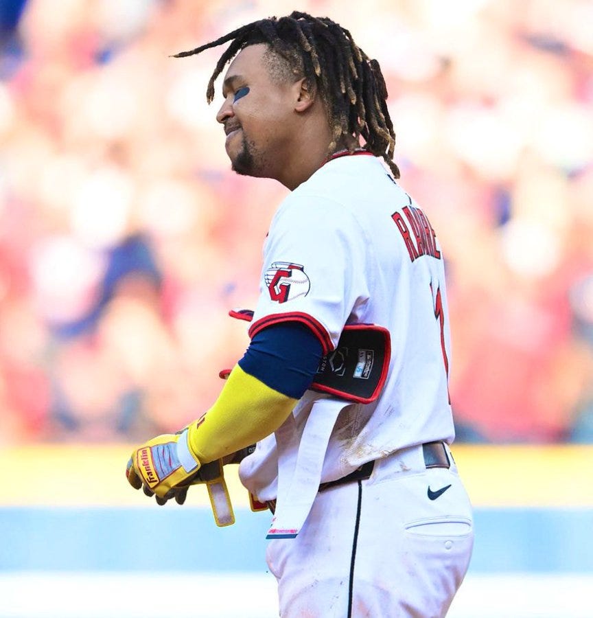 A baseball player wearing a white jersey with "Ramirez" and the number 11 on the back, along with yellow batting gloves and a black belt. The player has dreadlocks and is on a field with a blurred crowd in the background.