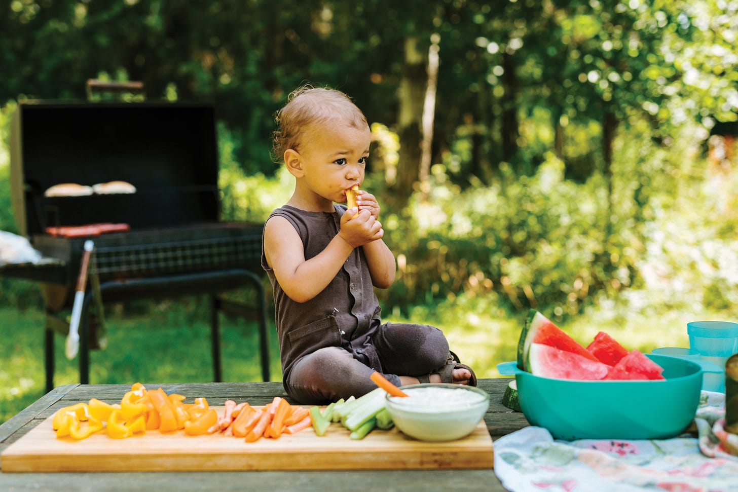 Child sitting on picnic table eating a carrot stick, with veggies and dip and cut watemelon on the table