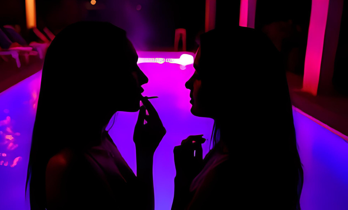 Two women sharing a cigarette at night by a pool, in conversation