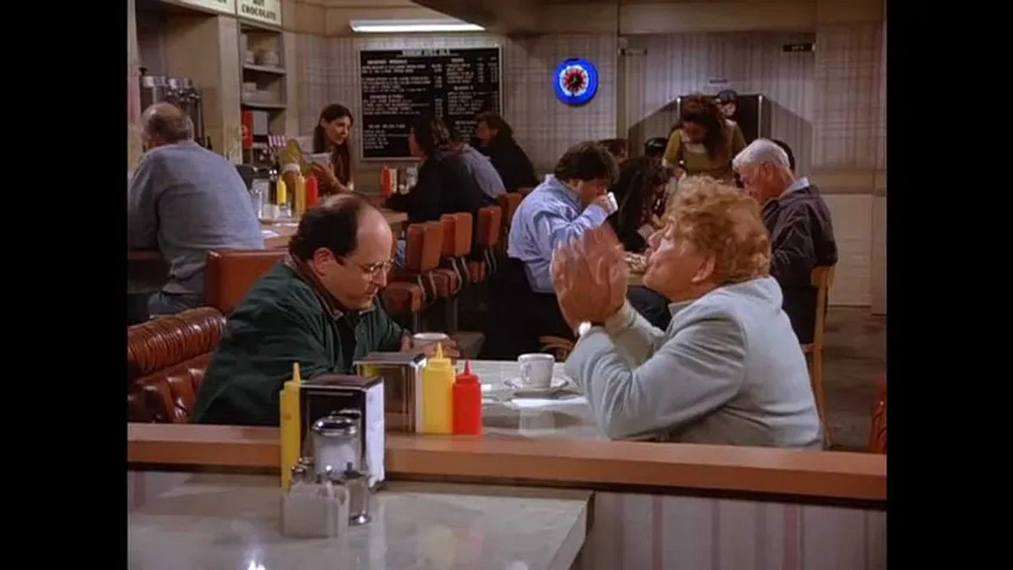 George Costanza sitting at the Monk’s Cafe counter, talking to one other person in a nearly empty diner. George Costanza sitting at the Monk’s Cafe counter, talking to one other person in a nearly empty diner.
