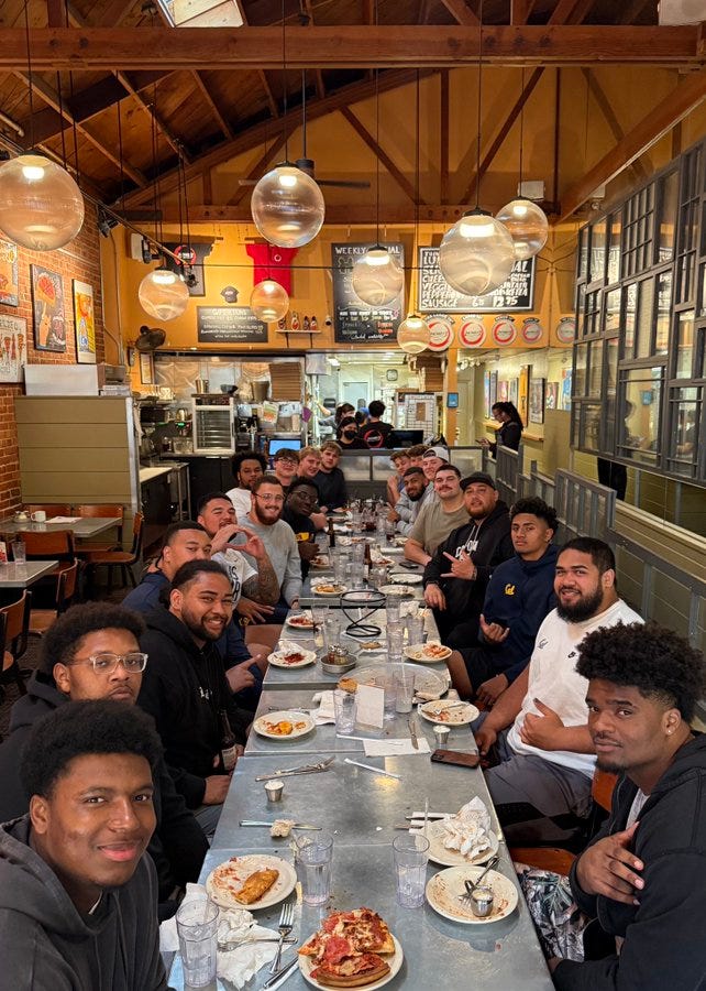 The image shows a large group of people, likely a sports team given the context of the post, gathered around a long table in a restaurant. The setting is Zachary's Pizza, as mentioned in the post, with a rustic interior featuring wooden beams and hanging lights. The table is filled with plates of pizza, drinks, and utensils, indicating a hearty meal has just been enjoyed. The atmosphere appears convivial and celebratory, with everyone seemingly content after their meal. The post text humorously notes that 'There was nothing left…' suggesting the group's large appetite, and tags the location with thanks to Zachary's Pizza. The image shows a large group of people, likely a sports team given the context of the post, gathered around a long table in a restaurant. The setting is Zachary's Pizza, as mentioned in the post, with a rustic interior featuring wooden beams and hanging lights. The table is filled with plates of pizza, drinks, and utensils, indicating a hearty meal has just been enjoyed. The atmosphere appears convivial and celebratory, with everyone seemingly content after their meal. The post text humorously notes that 'There was nothing left…' suggesting the group's large appetite, and tags the location with thanks to Zachary's Pizza.