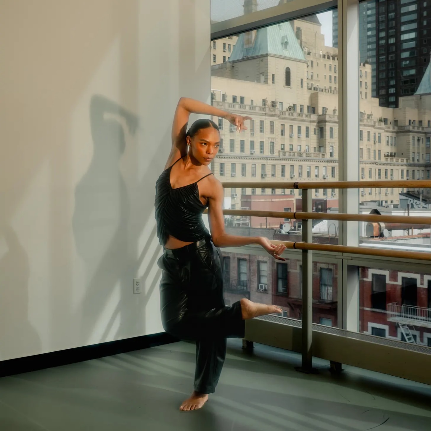 A photo of a young Black femme dancing in a studio with floor to ceiling windows with sweeping views of Manhattan.